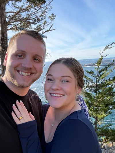 Tim and Elise Morgan smiling together on a rocky coastline, with pine trees and the ocean behind them