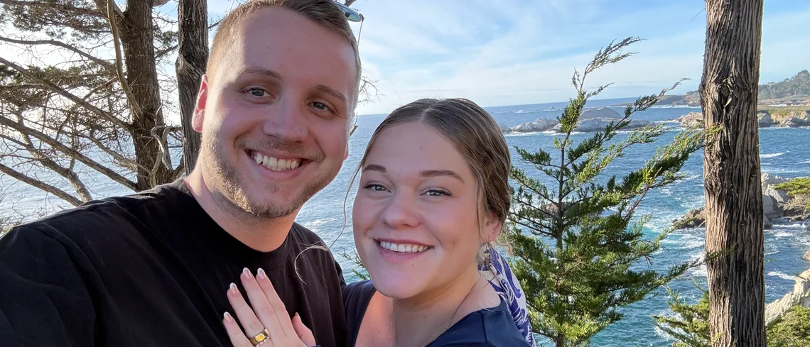Tim and Elise Morgan smiling together in a selfie on a rocky coast, with pine trees and the ocean behind them