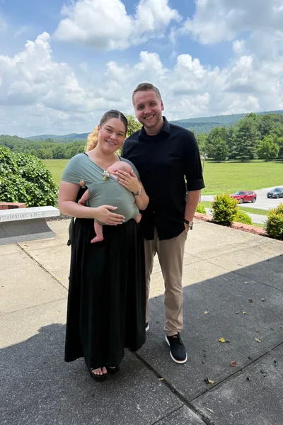 Tim and Elise Morgan with their baby in a carrier, standing outdoors on a sunny day with green hills behind them